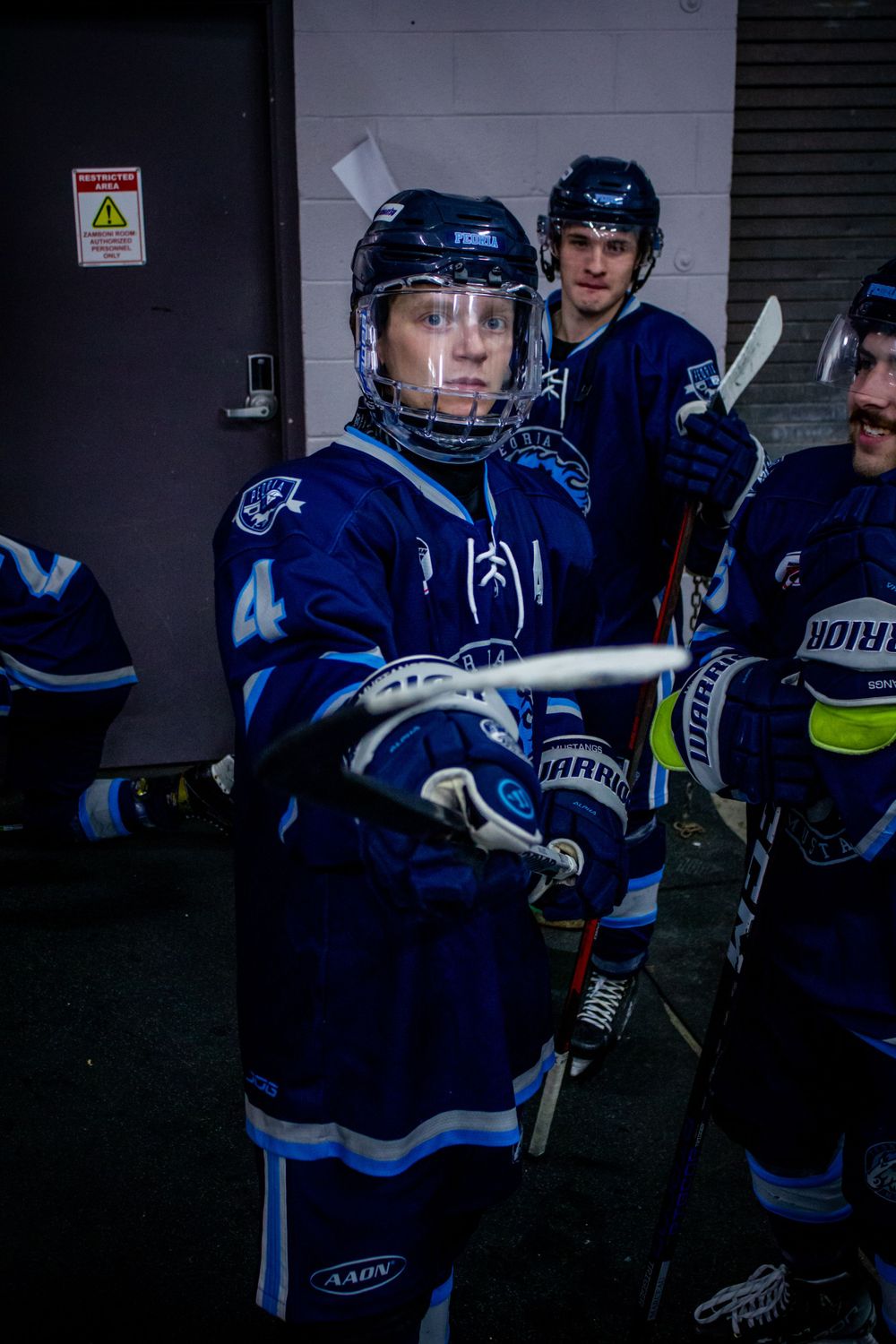 Peoria Mustangs Hockey player Declan McDermott in dark blue jersey with number 4 stands ready in corridor.