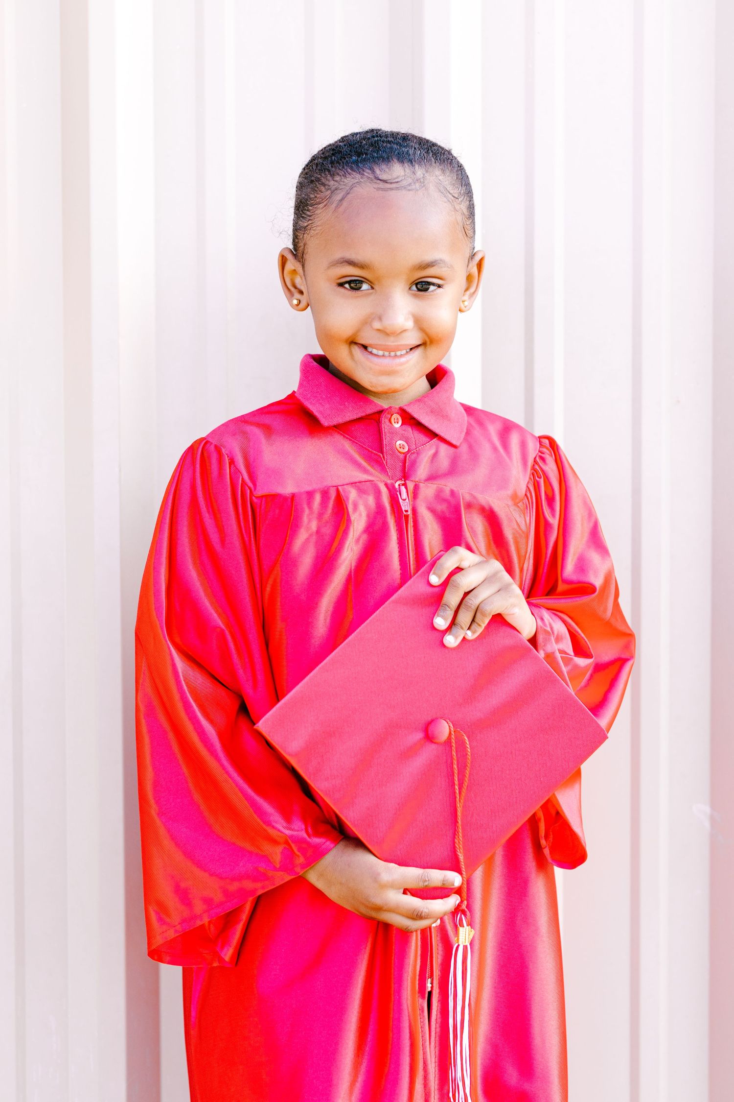 Young student wearing bright red graduation gown holds matching diploma cover while smiling against white background.