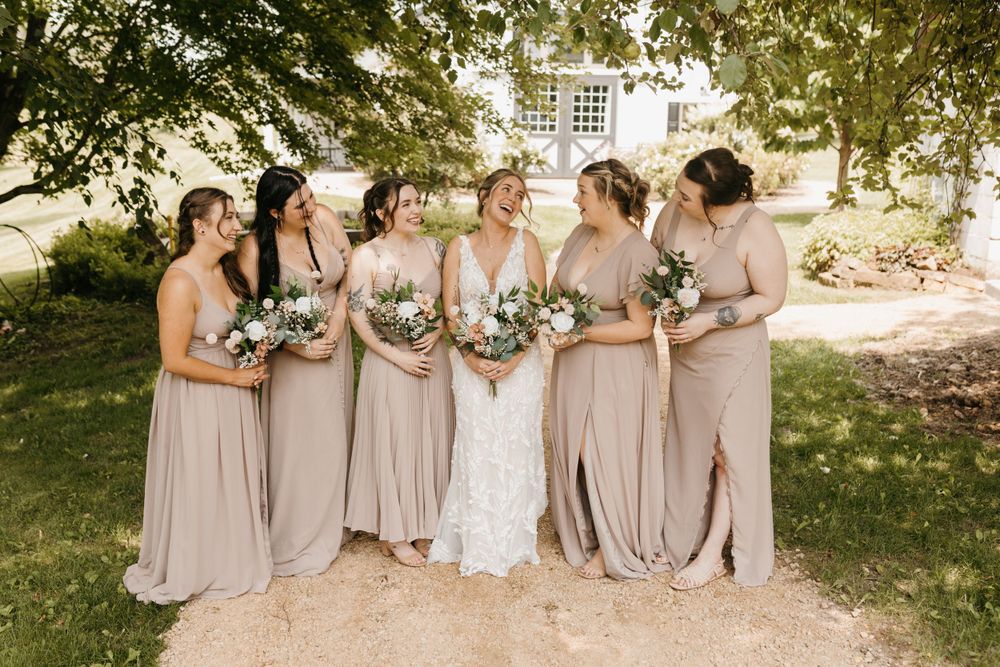 Bride and bridesmaids in taupe gowns laugh together outdoors holding white and green bouquets.