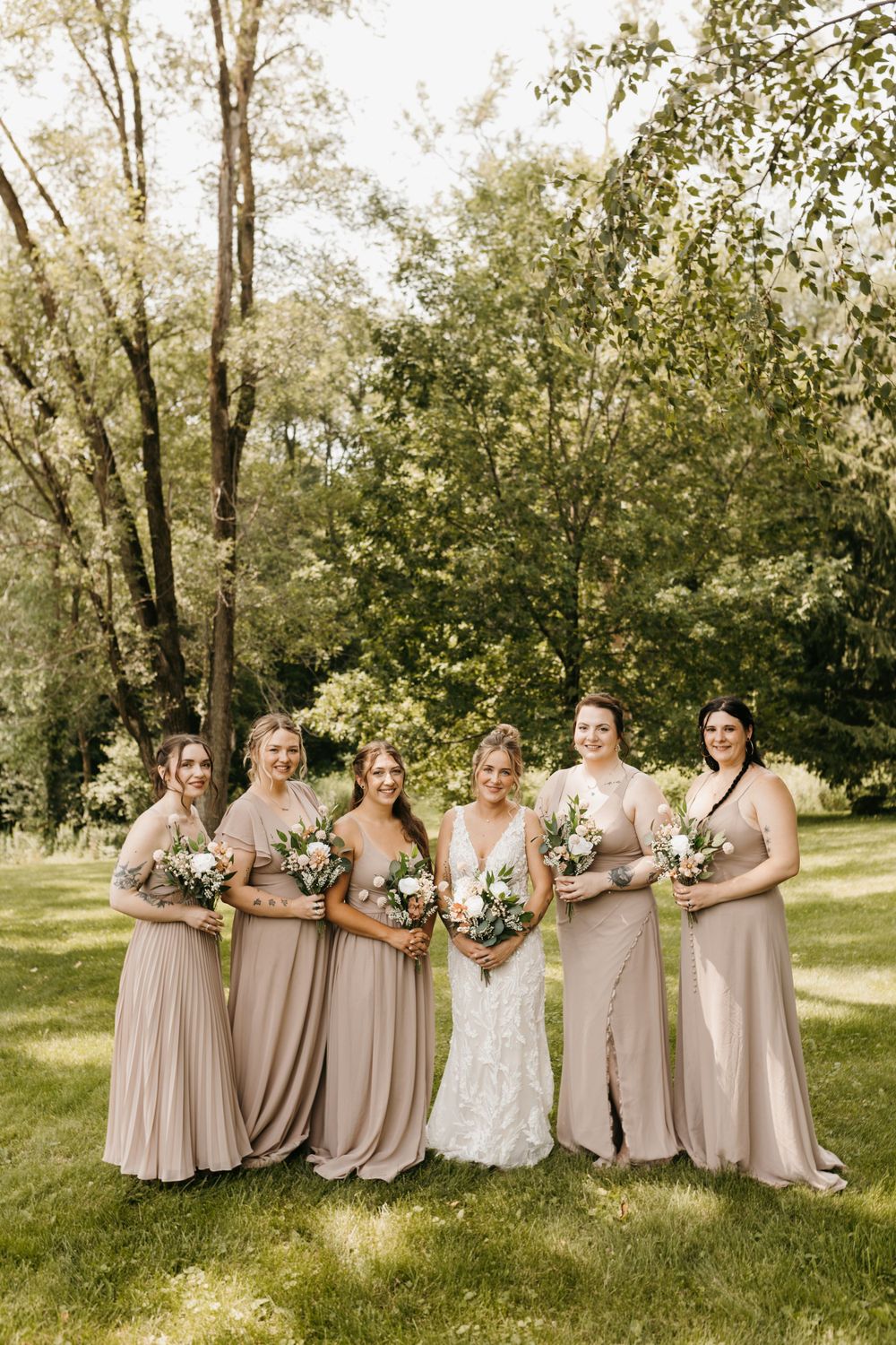 Bride in lace gown stands with five bridesmaids in taupe dresses on a sunny green lawn.
