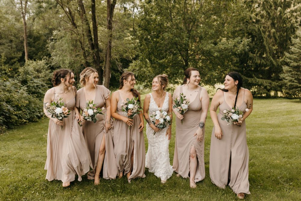 Bride and bridesmaids in taupe gowns walk and laugh together across a lush green lawn.