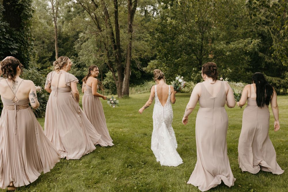 Bride in backless white gown walks with bridesmaids in taupe dresses across a green lawn.