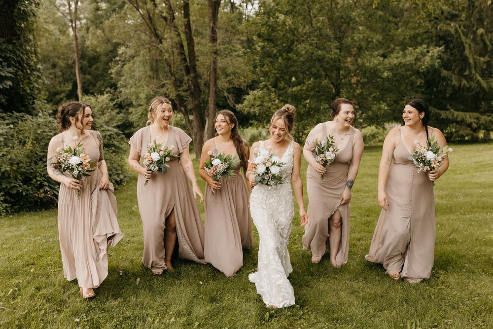Bride and five bridesmaids in taupe gowns walk together laughing on a green lawn outdoors.