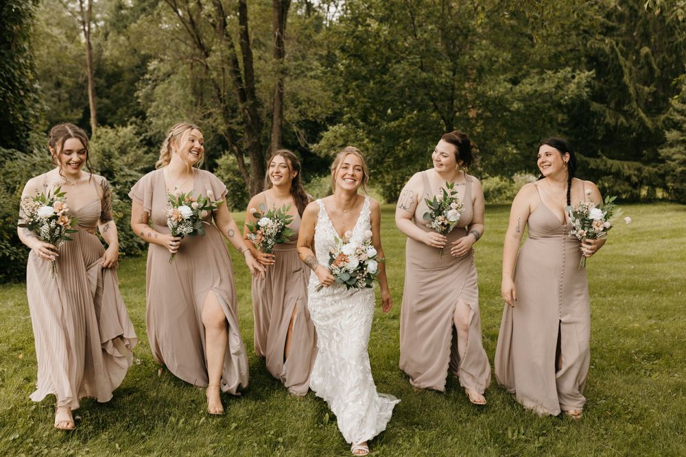 Bride in lace gown laughs with bridesmaids in taupe dresses holding bouquets on a green lawn.