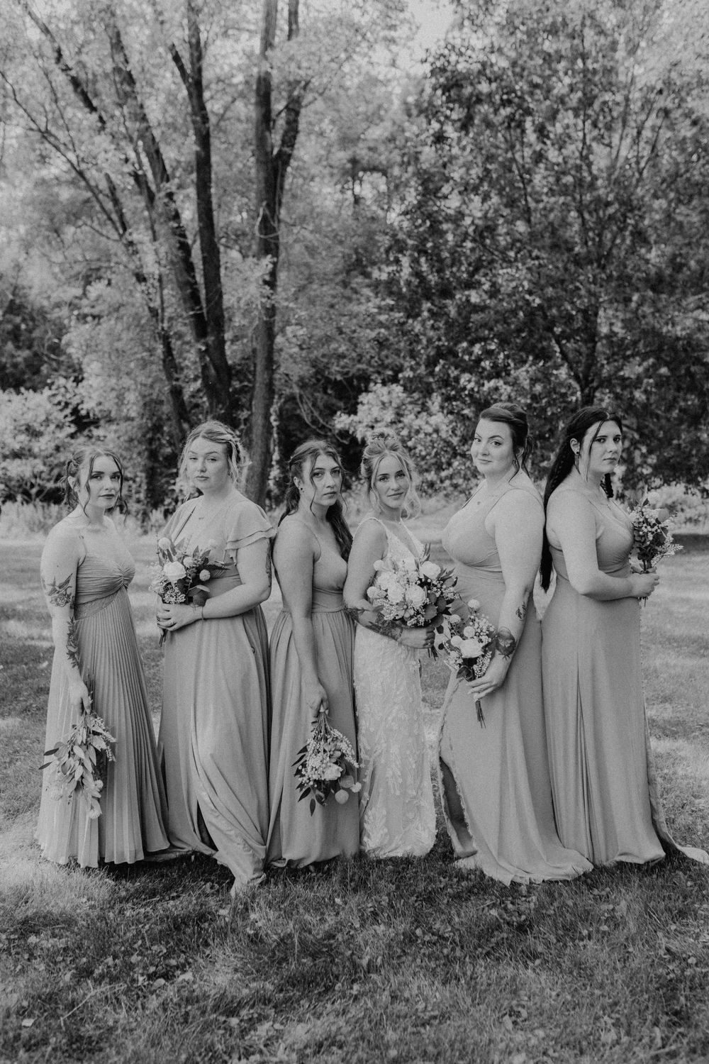 Black and white photo of bride and five bridesmaids in long gowns posing outdoors among trees.