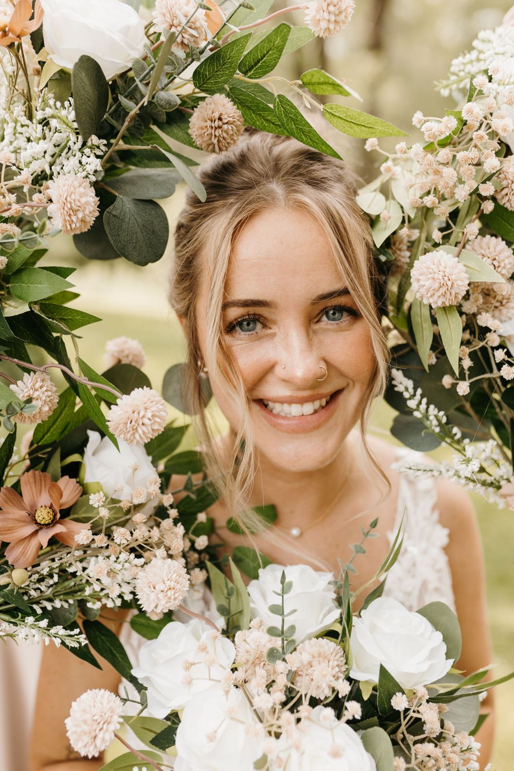 Smiling bride with blue eyes surrounded by lush white and blush wedding bouquet with greenery.