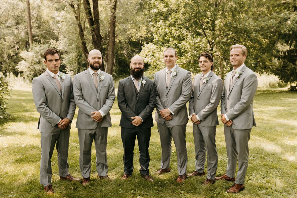Groom in dark suit stands with five groomsmen in gray suits outdoors on green lawn.
