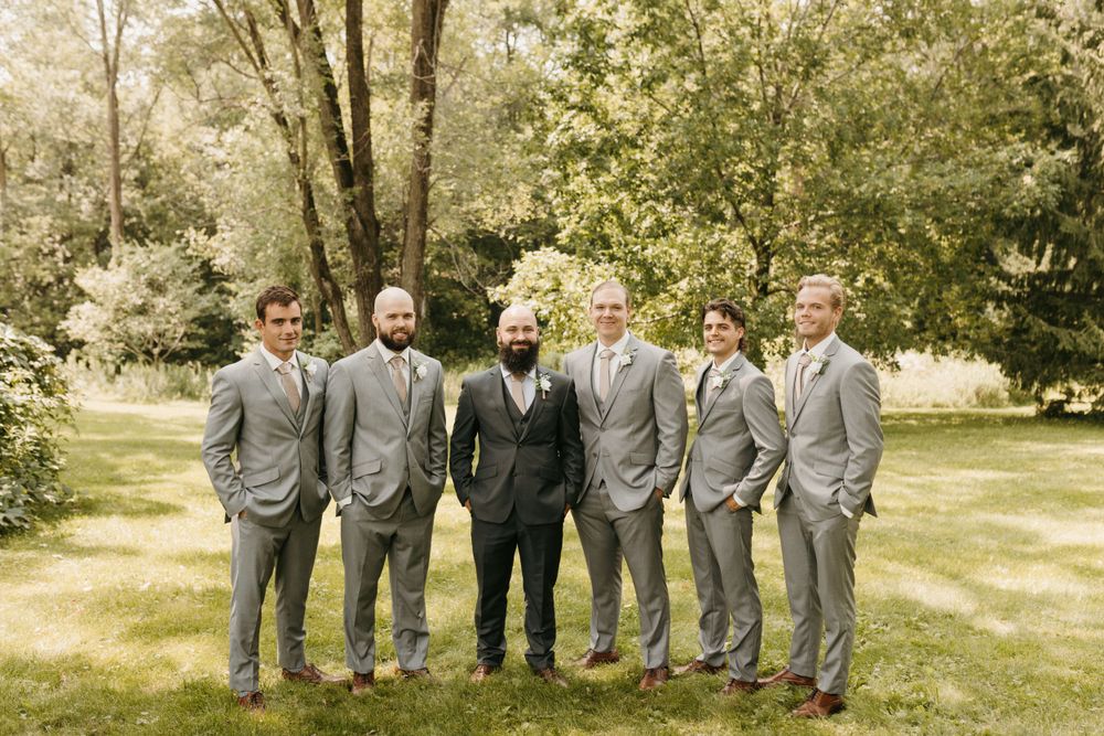 Groom and groomsmen in gray suits smiling together in a sunny outdoor garden setting.