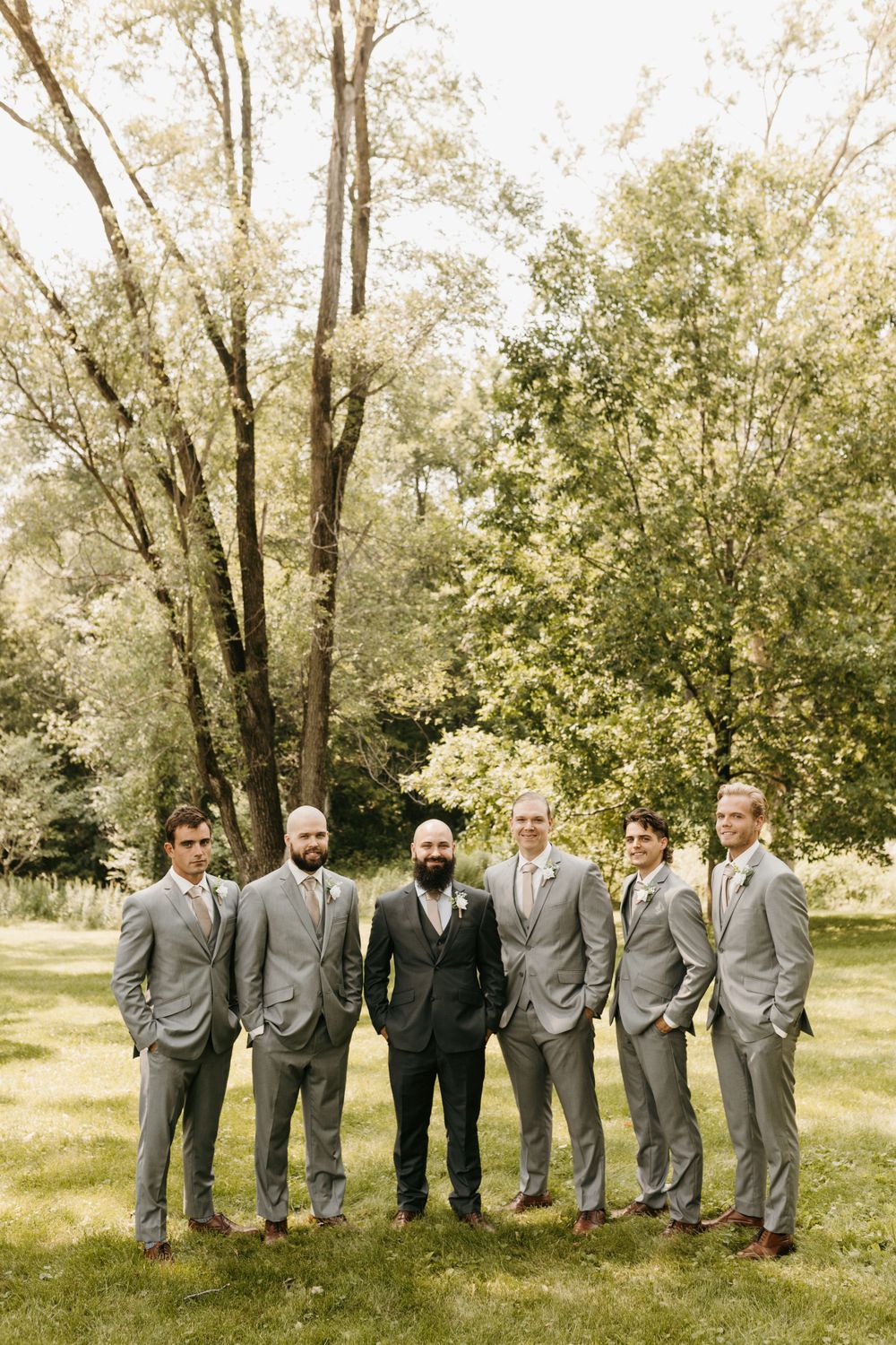 Groom in dark suit poses with five groomsmen in gray suits among tall trees outdoors.