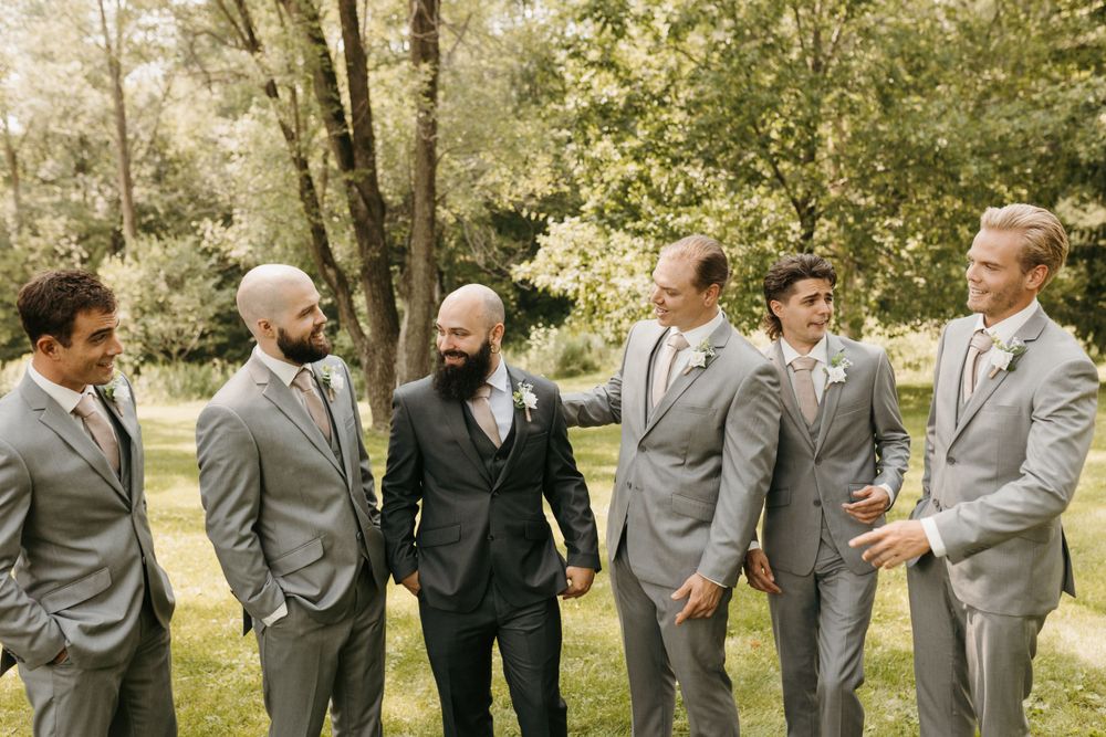 Groom and groomsmen in gray suits laughing together in a lush outdoor wedding setting.