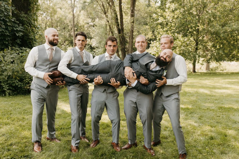 Five groomsmen in gray suits playfully lifting the groom horizontally outdoors.