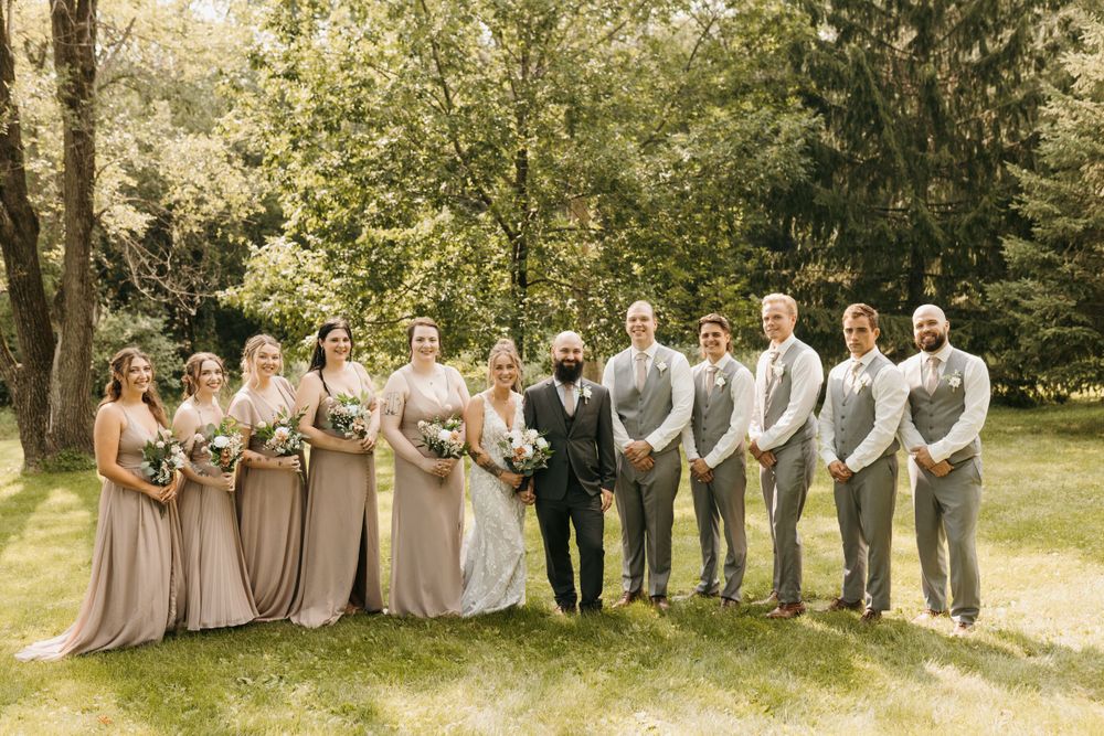 Full wedding party in blush bridesmaid dresses and gray groomsmen suits posing outdoors.
