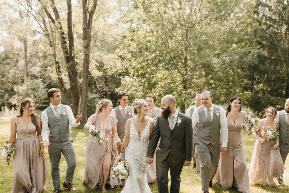 Wedding party walks outdoors in gray suits and taupe bridesmaid dresses amid lush green trees.