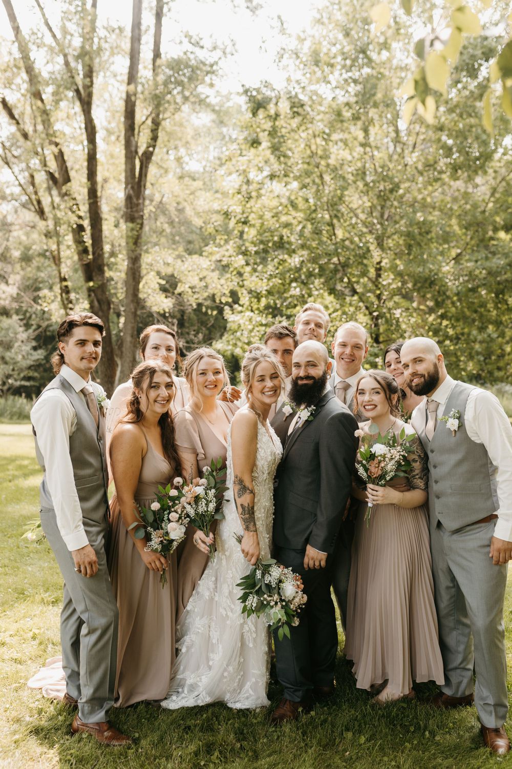 Smiling wedding party poses together outdoors with bride and bearded groom at center.