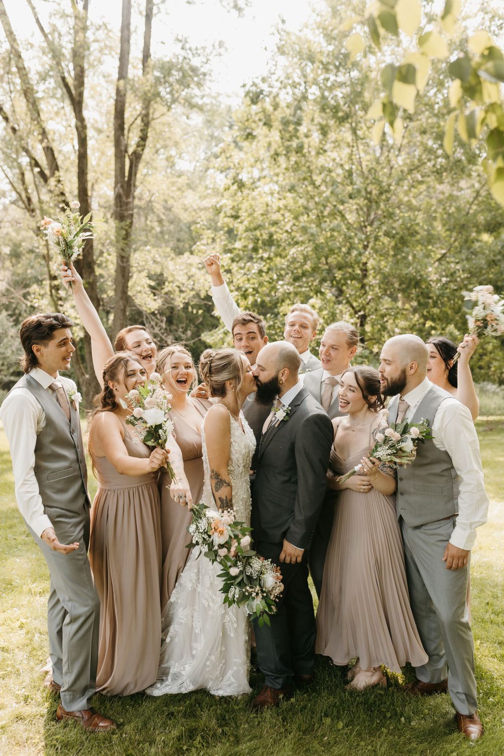 Wedding party cheers as bride and bearded groom share a kiss outdoors under green trees.