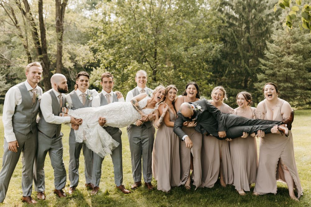 Laughing wedding party lifts bride and groom horizontally outdoors on green grass.