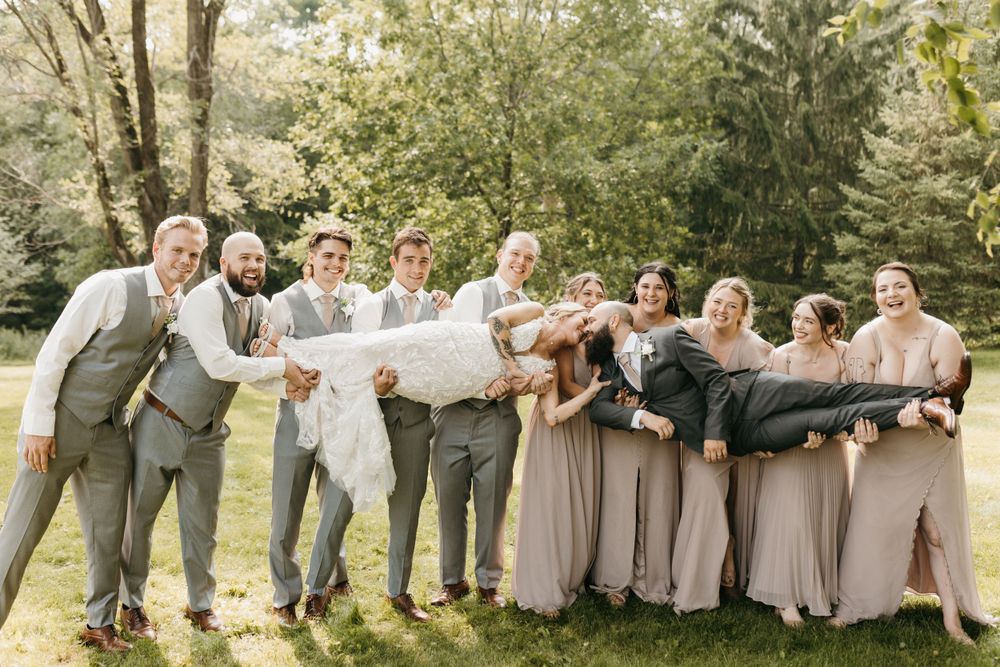 Bridal party playfully carries bride and groom horizontally while laughing outdoors.