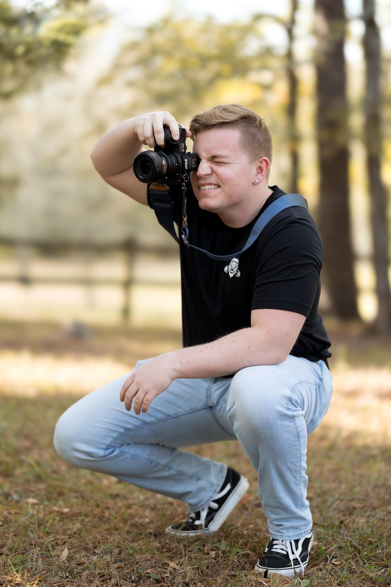 A young male photographer crouches outdoors while taking a photo with a DSLR camera.