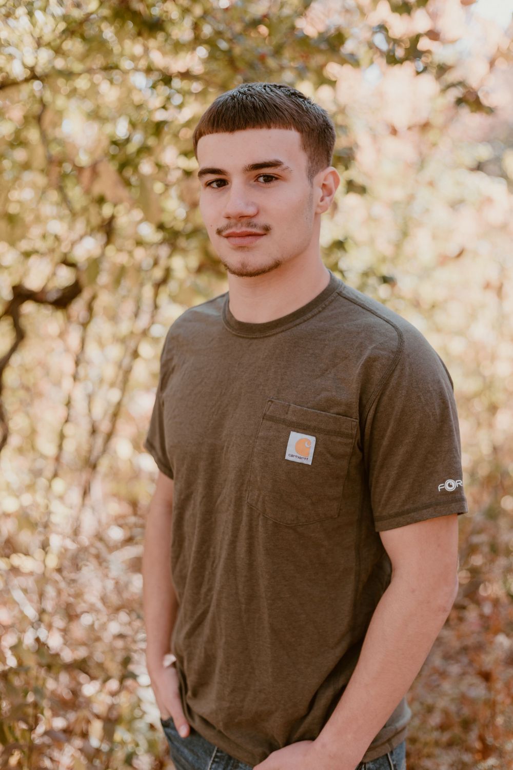 Young man in olive Carhartt t-shirt standing outdoors among golden autumn foliage.