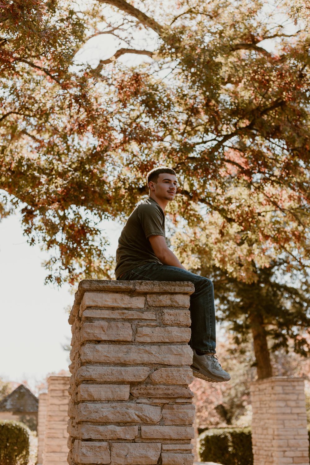 Young man sitting atop a stone pillar under a colorful autumn tree outdoors.