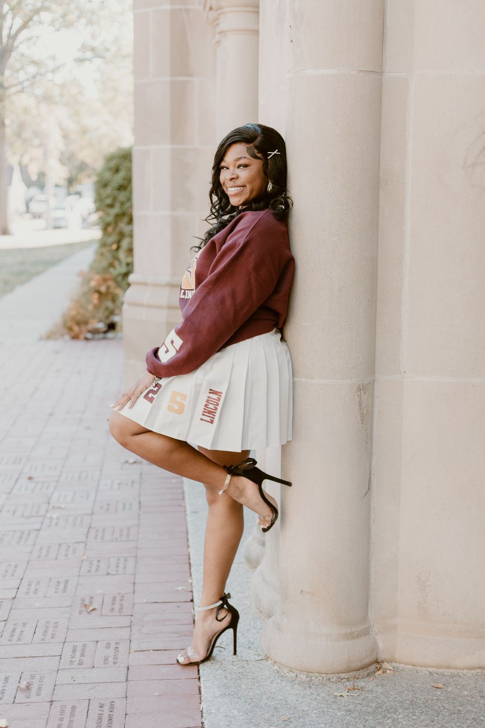 Young Black woman in maroon jacket and pleated skirt leaning against stone column outdoors.