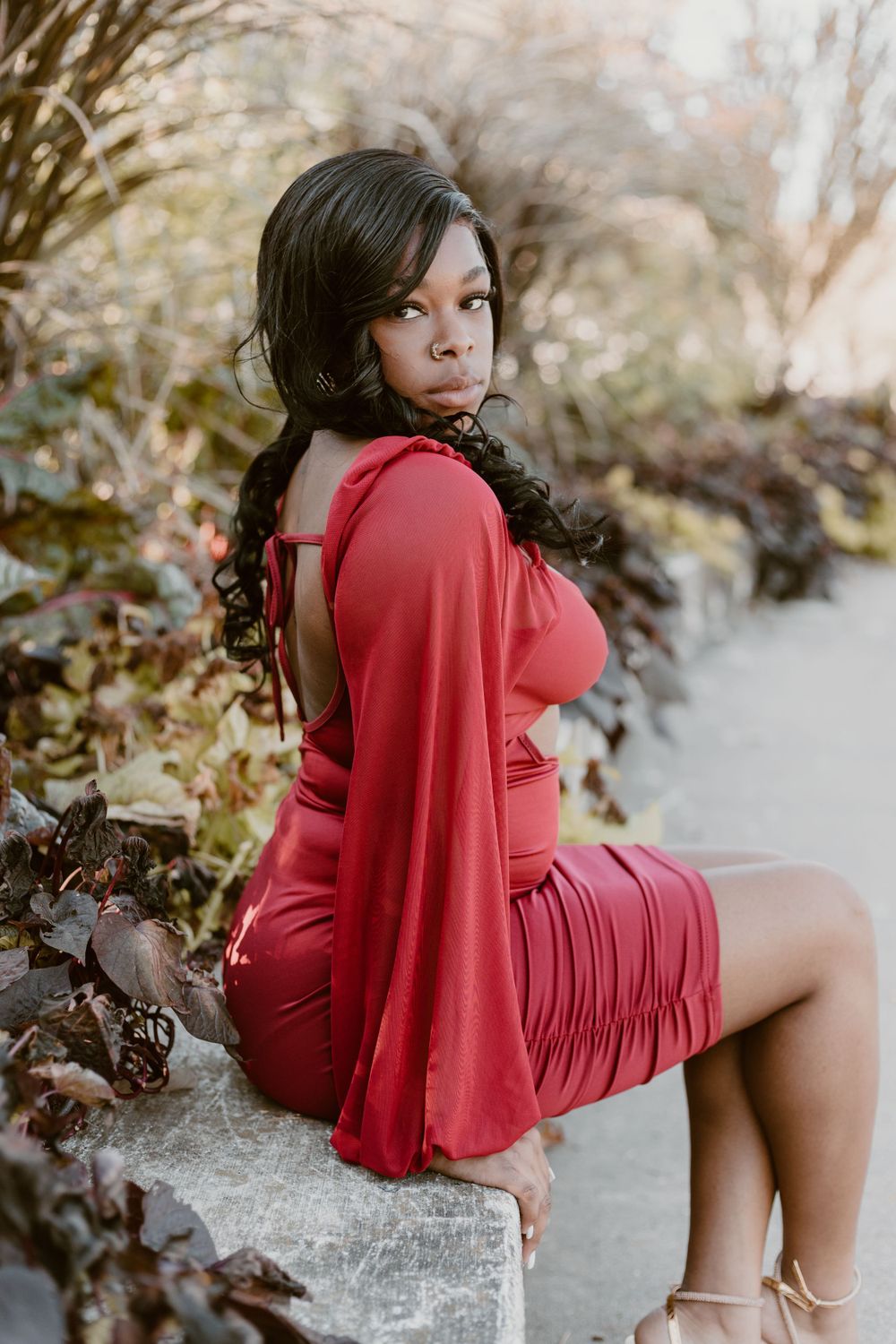 Young Black woman in elegant red backless dress sitting on stone ledge in garden.