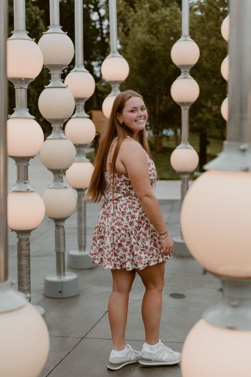 Smiling young woman in floral mini dress posing among glowing globe lights at outdoor installation.