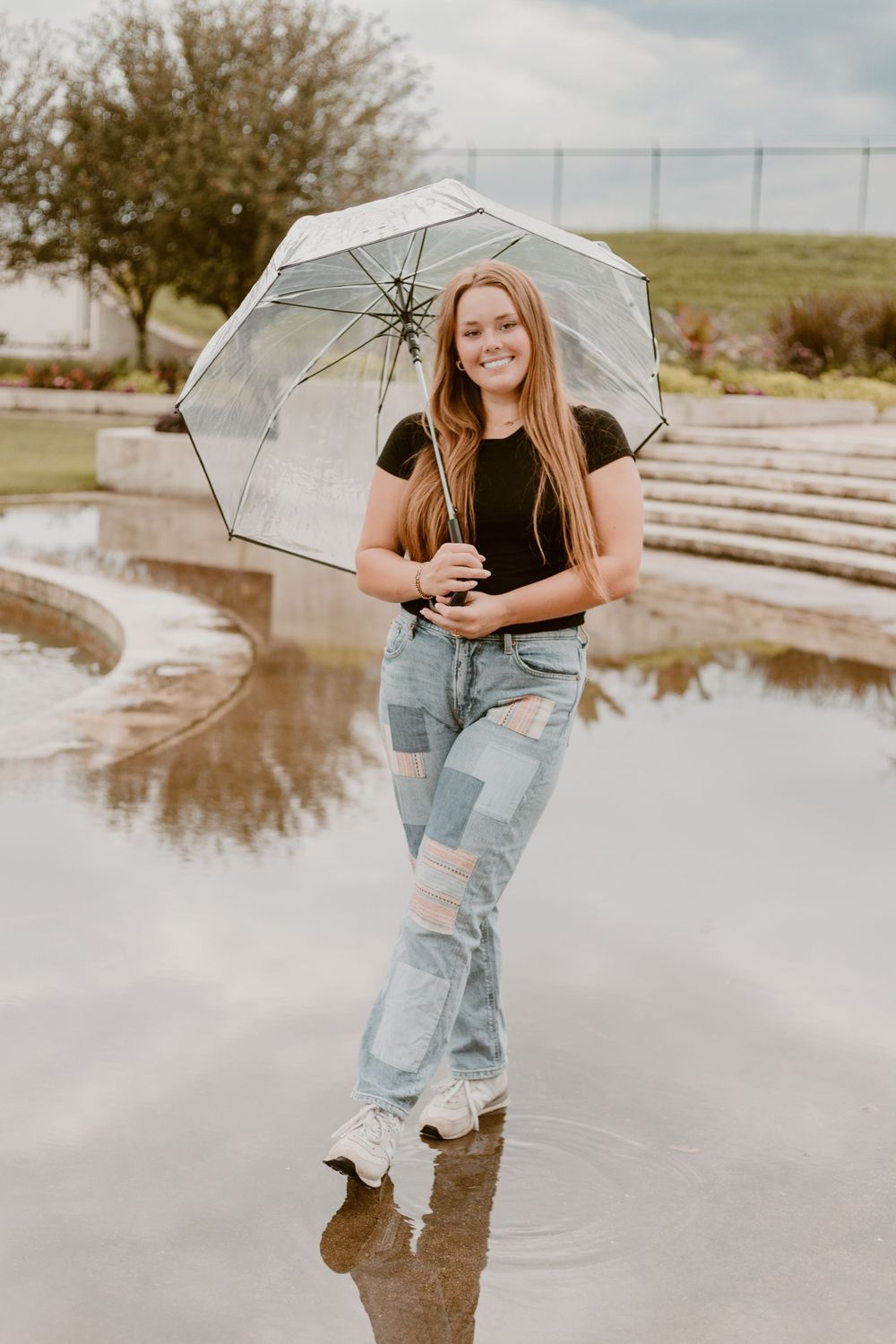 Smiling teenage girl holding clear umbrella standing in rain puddle wearing patchwork jeans and black top.
