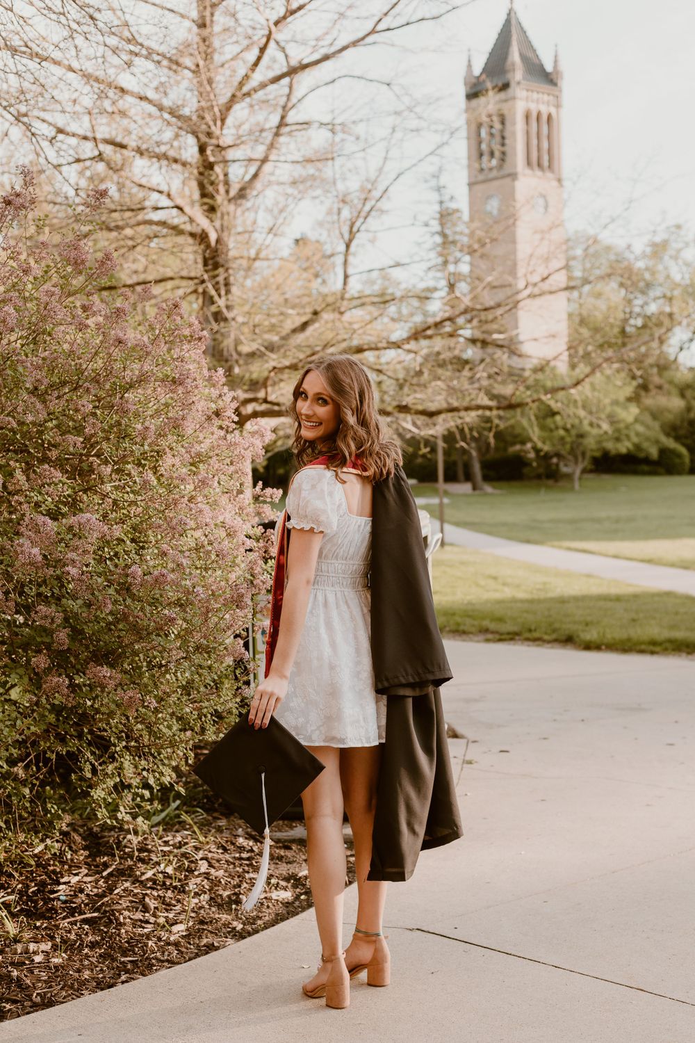 College graduate in white dress holding cap and gown walking near flowering trees and campus bell tower.