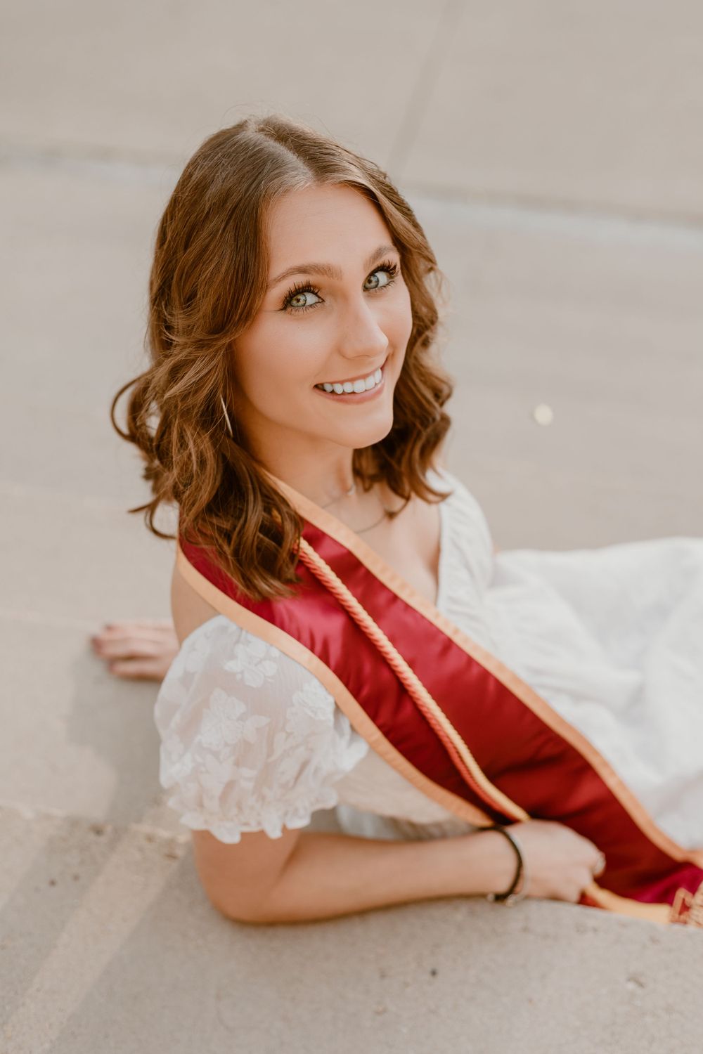 Smiling young woman in white dress with red graduation sash lying on pavement looking up at camera.
