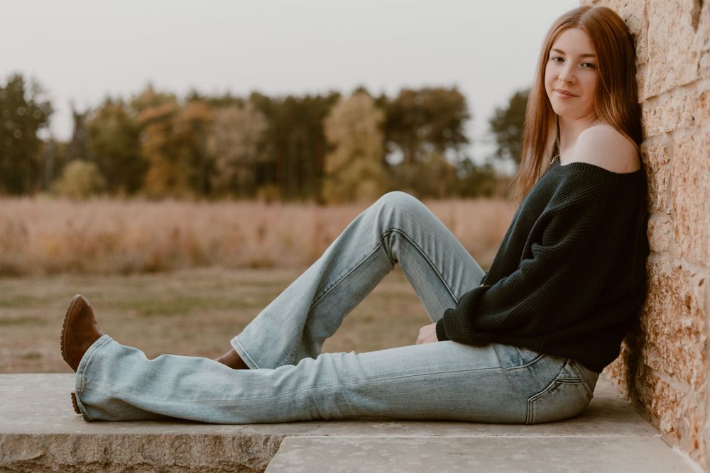Young woman in black off-shoulder sweater and jeans sitting against a rustic stone wall in an autumn field.