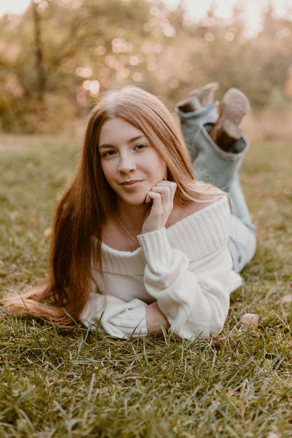 Teen girl with long red hair lying on grass in white off-shoulder sweater at sunset.
