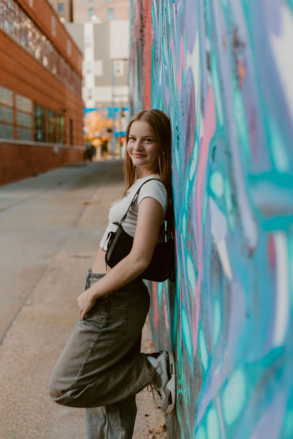 Teen girl smiling while leaning against colorful graffiti wall in urban alleyway.