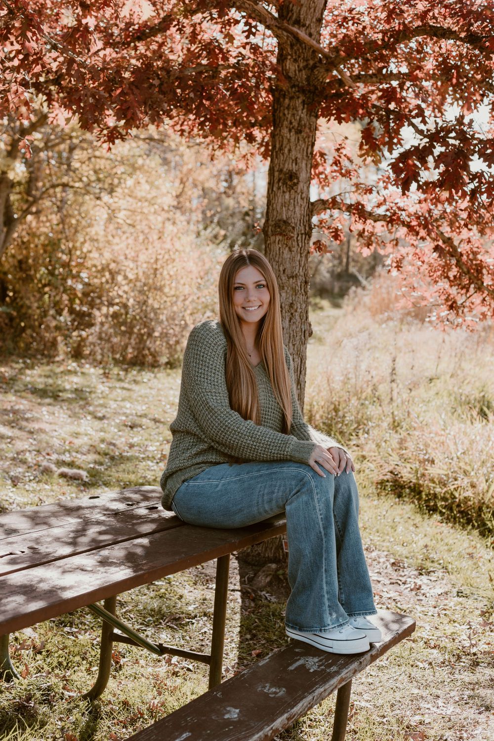 Teen girl in green sweater and jeans sitting on picnic table under red autumn tree.