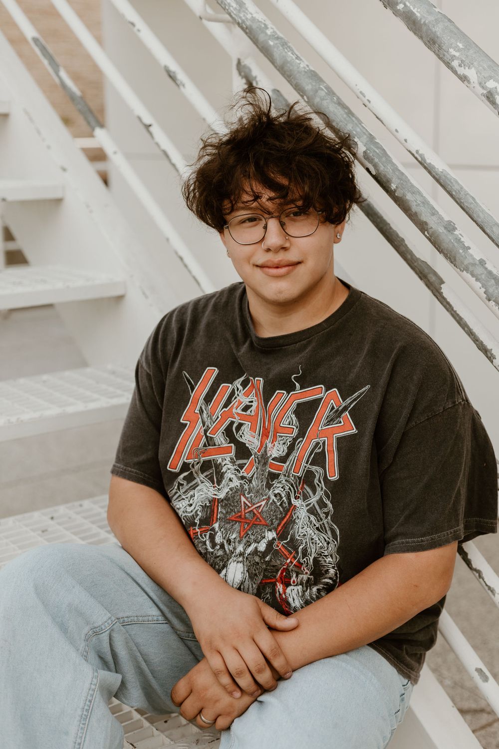 Teenage boy in Slayer graphic tee and glasses sitting on bleacher steps smiling at camera.