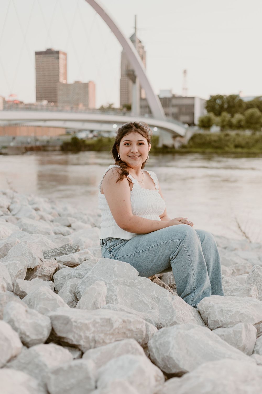 Young woman in white top and jeans sitting on rocks by river with city bridge skyline backdrop.