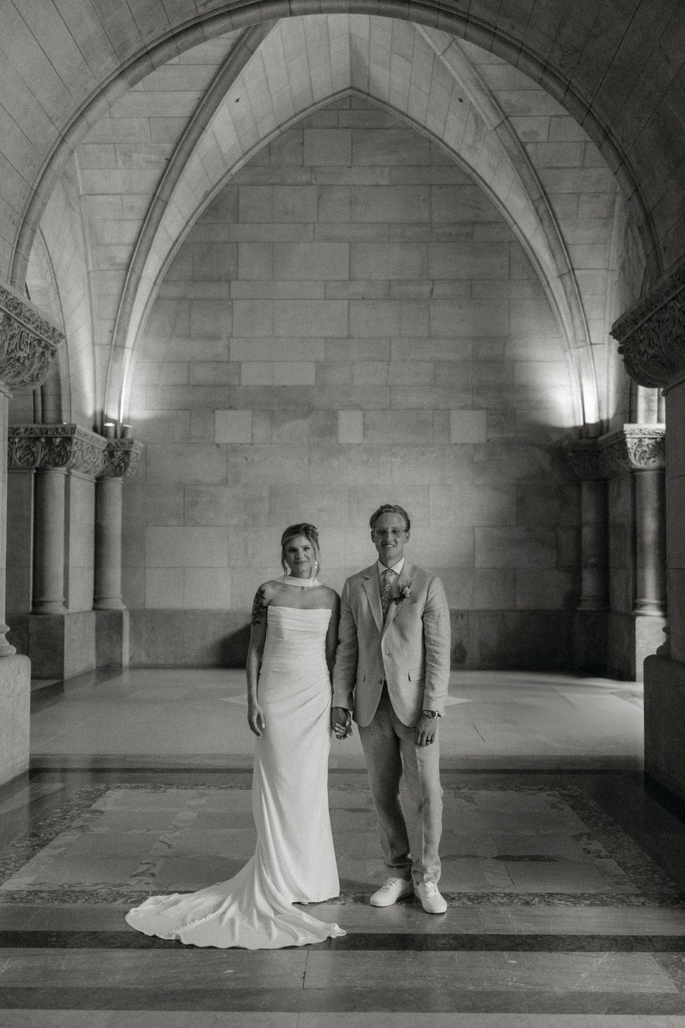 Black and white photograph of a couple standing in a grand arched stone hallway.