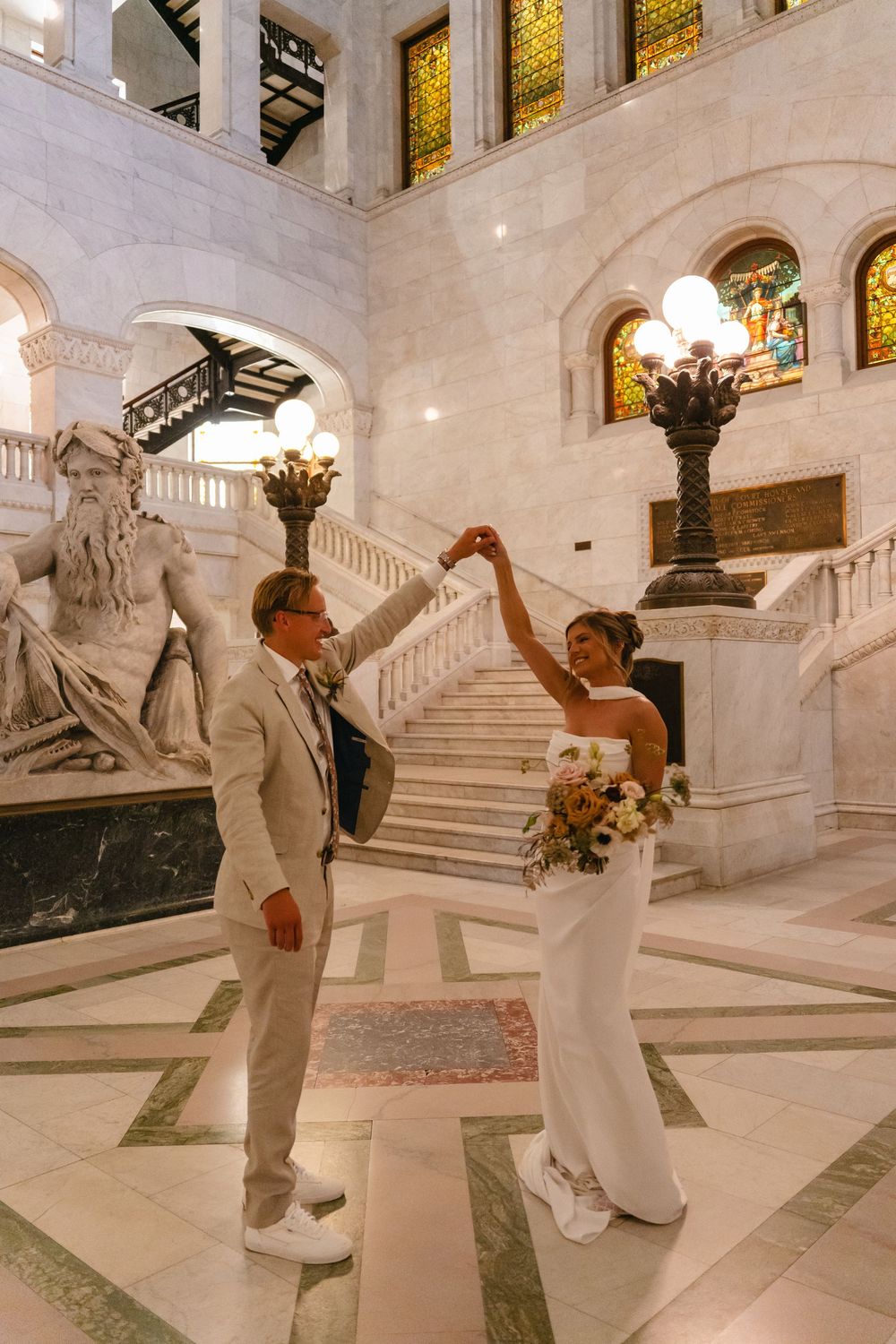 A wedding couple dances and twirls together on a grand marble staircase in an elegant historic building interior.