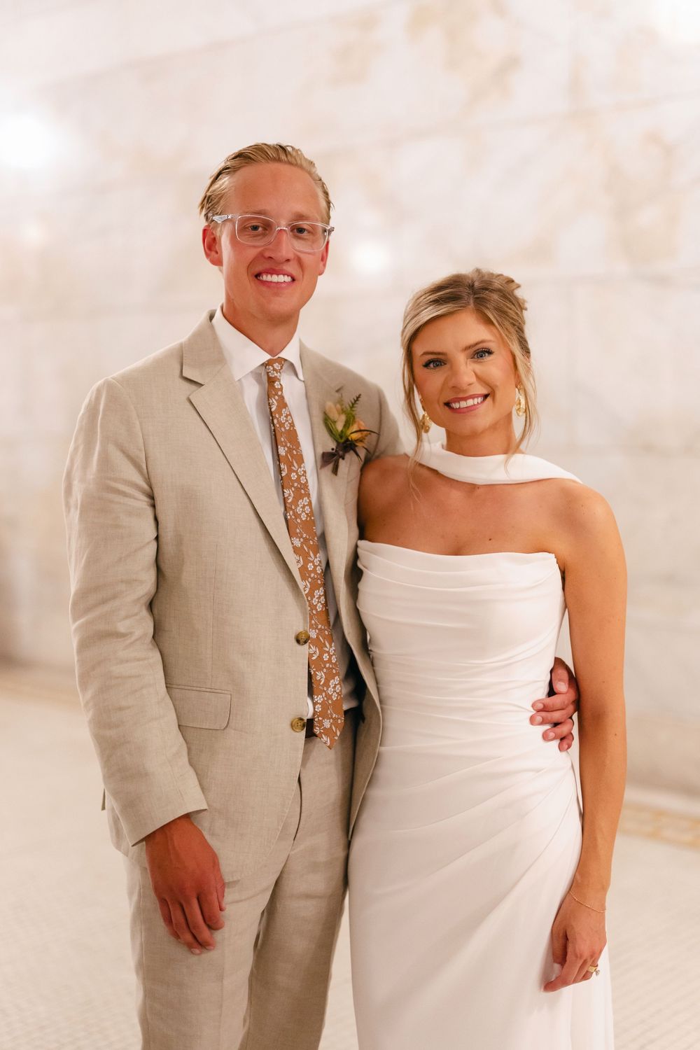Wedding couple poses together on beach in tan suit and white dress against soft beige background.