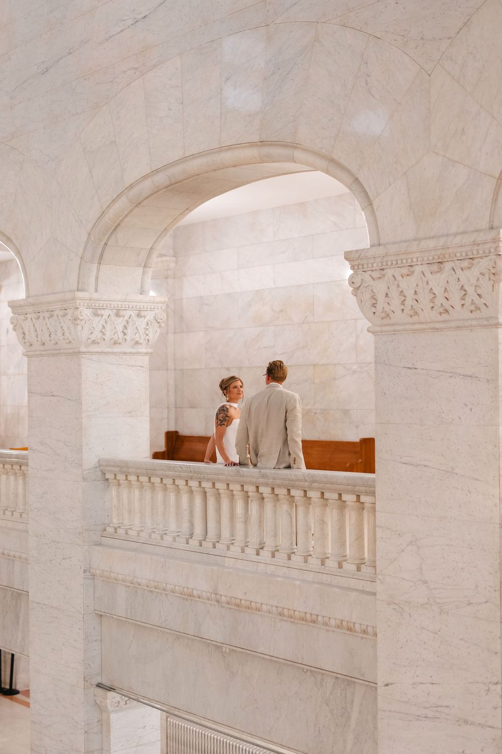 Two people stand on a white marble balcony with ornate architectural details in an elegant interior space.
