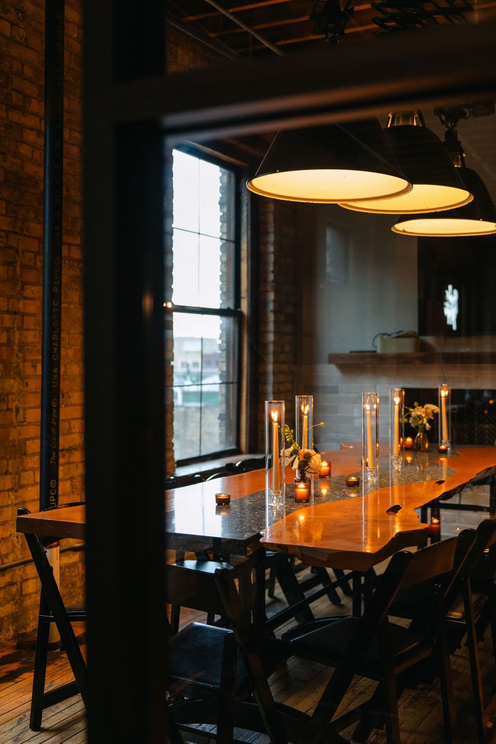Rustic restaurant dining room with wooden table, black chairs, and pendant lights against exposed brick wall.
