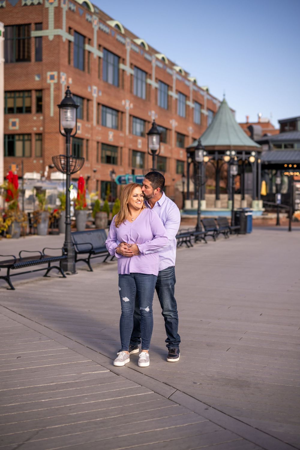 Couple embracing on boardwalk with brick building and decorative lampposts in background.