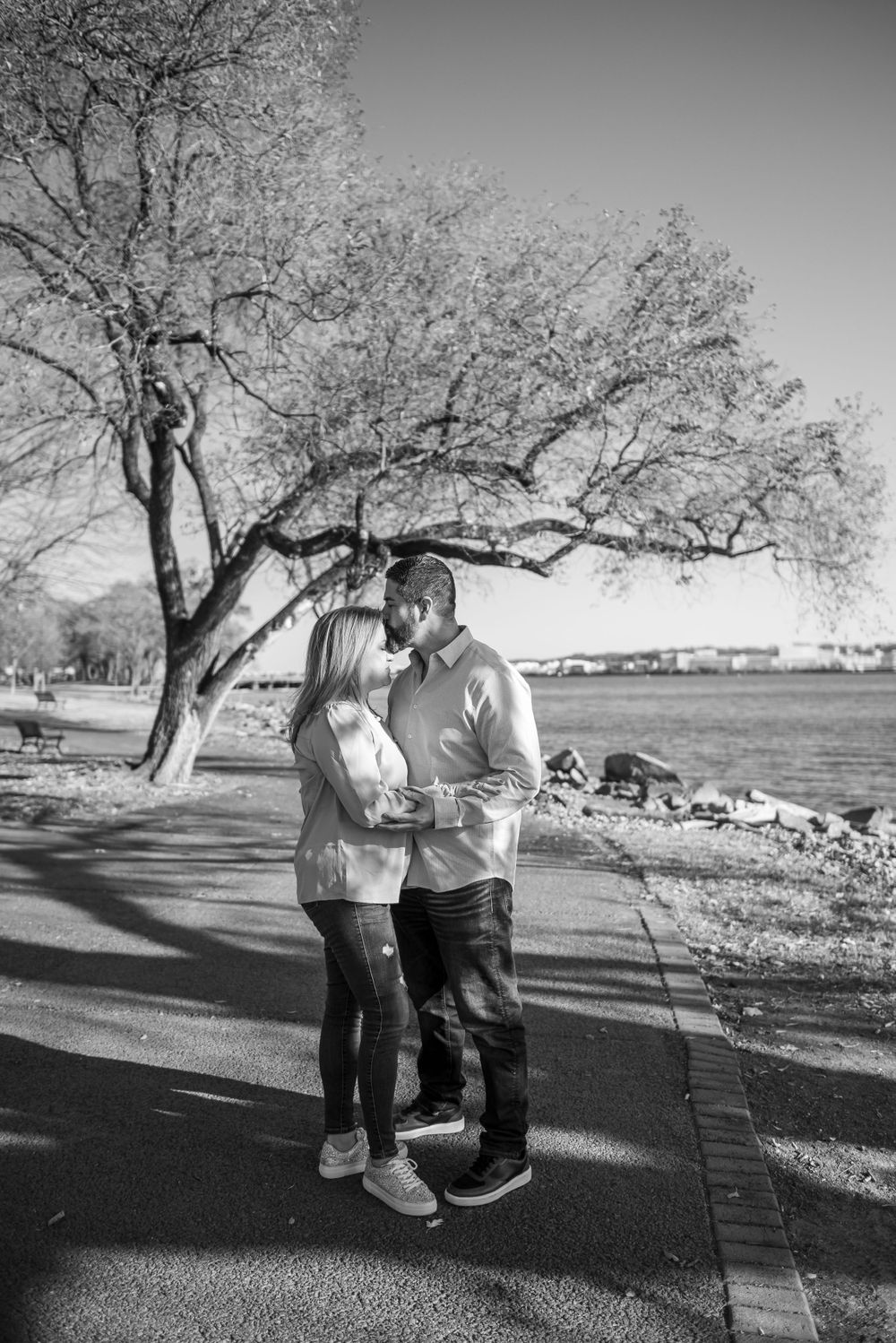 Romantic couple embraces under a blooming tree on a sunny path by the waterfront.