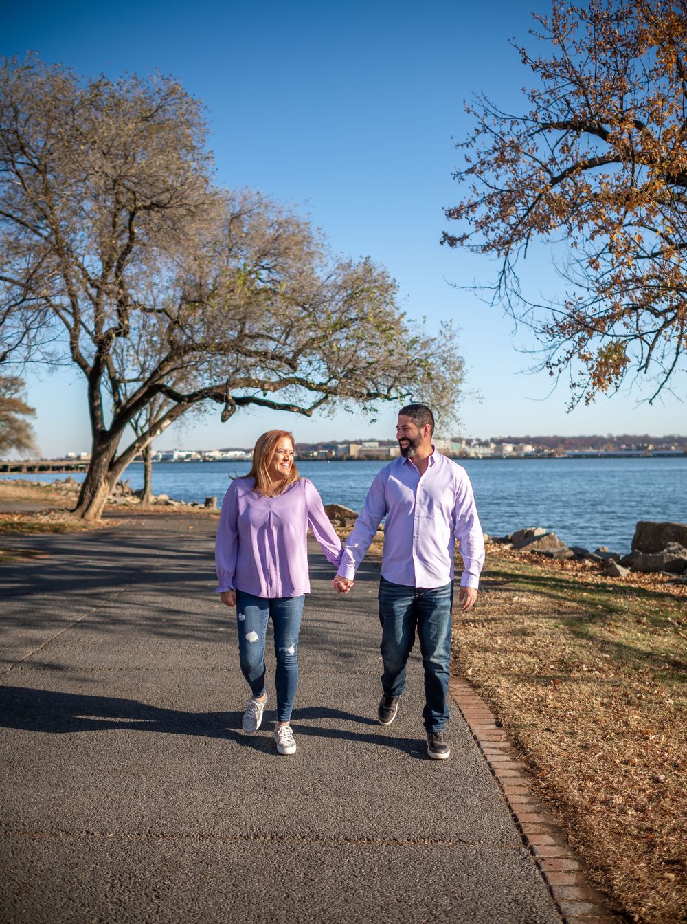 Couple walking hand in hand along waterfront path with trees and blue sky overhead.