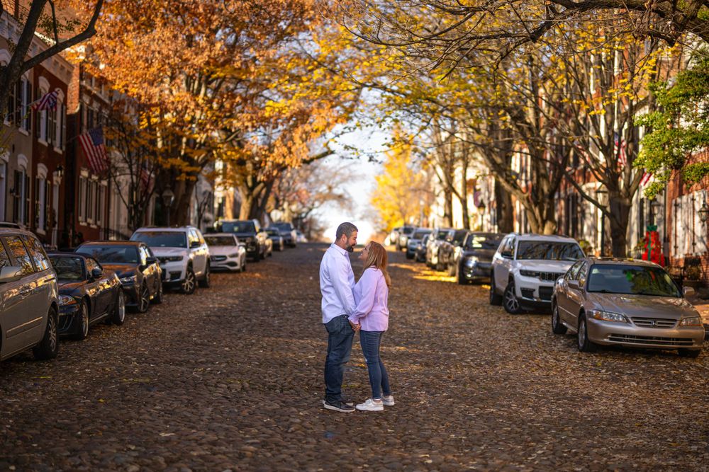 Couple stands together on autumn street with golden trees and parked cars.