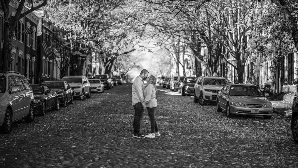 Black and white photo of couple embracing on leaf-covered street lined with parked cars.