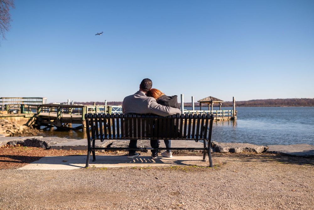 A couple sits together on a bench overlooking a scenic waterfront dock area on a sunny day.