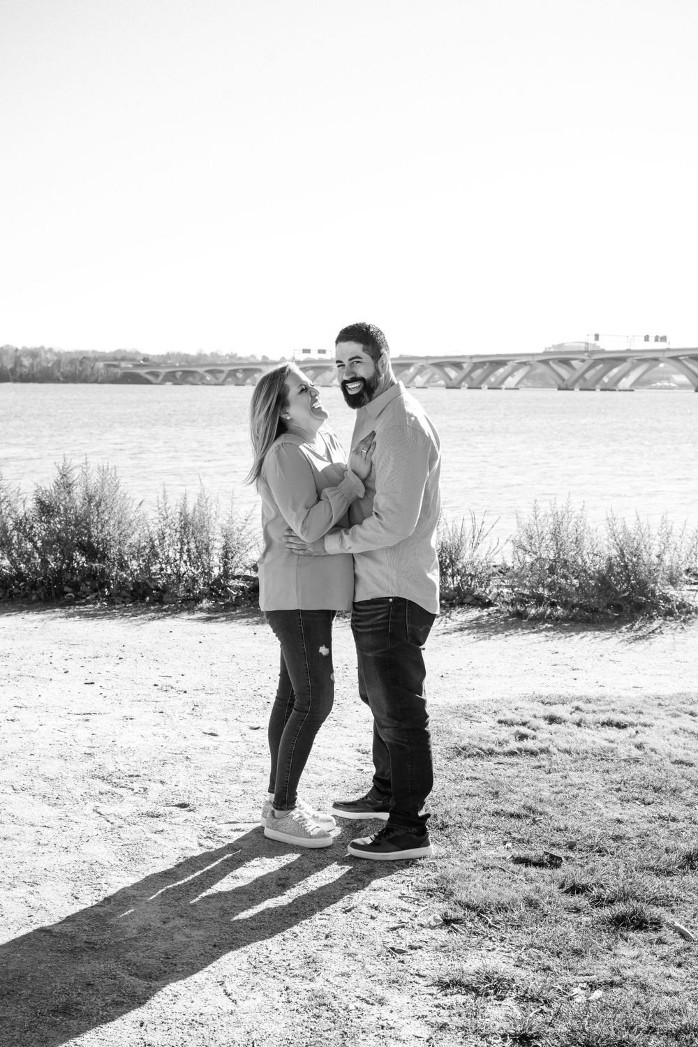 A romantic black and white portrait of a couple embracing by a riverside with a bridge in the background.