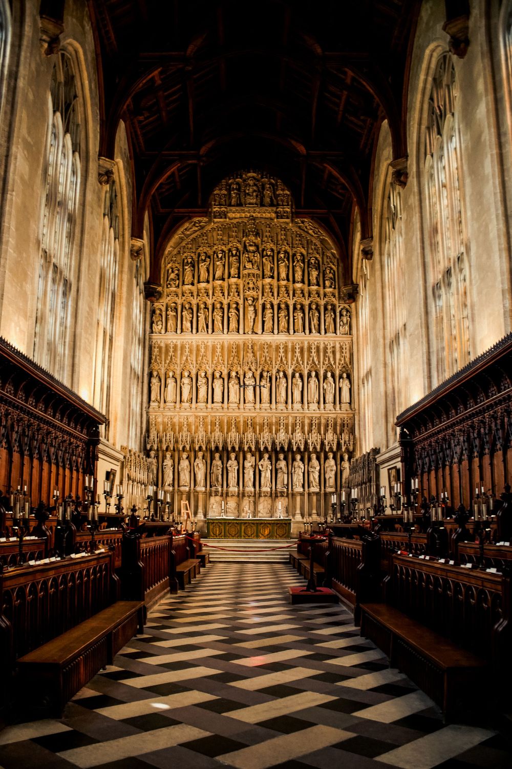 Interior view of a Gothic cathedral with ornate reredos and checkerboard flooring leading to an elaborate golden altar screen.