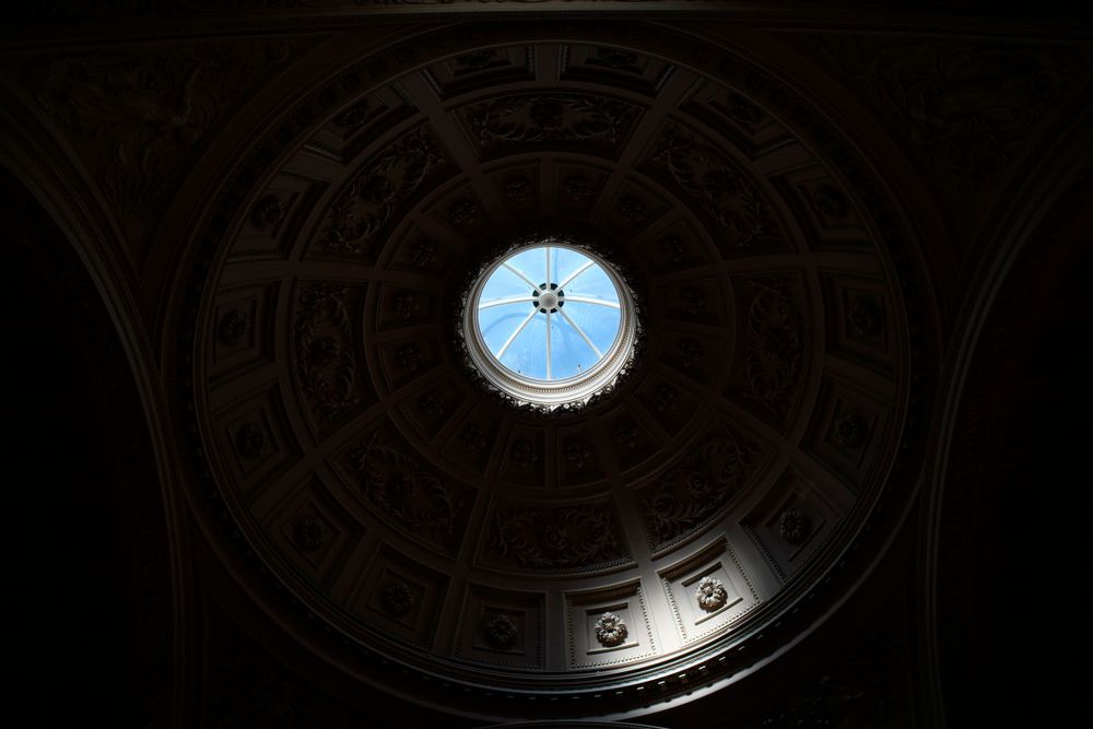 Circular skylight opening in an ornate domed ceiling revealing blue sky.